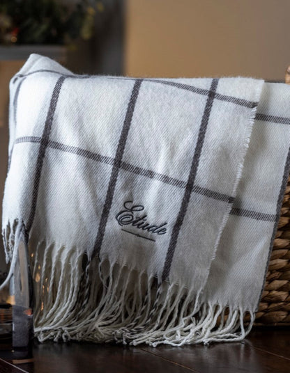 White plaid throw blanket with fringe and "Etude" embroidered on it, draped in a basket.